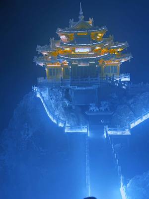 A nighttime shot of a multi-tiered, illuminated traditional Asian building perched atop a rocky mountain. The structure is intricately designed and bathed in warm yellow light, contrasting against the deep blue hues of the night sky and surroundings. Steps lead up to the building, and the entire scene evokes a mystical or spiritual atmosphere.
