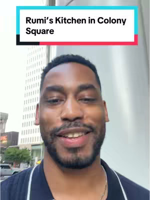 A man with a beard and a dark blue shirt is smiling outside with a city in the background. Text overlay at the top says 'Rumi's Kitchen in Colony Square'.