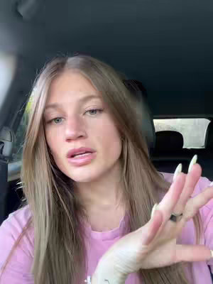 A woman with long blonde hair is sitting in a car. She is wearing a pink shirt and has her hand up, as if she is talking. The background shows the inside of a car with seats and a window.