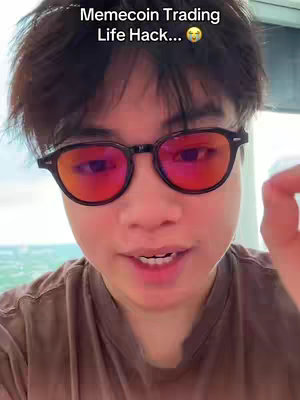 A selfie-style video cover image featuring a young man with dark hair and glasses. The glasses have red-tinted lenses, with the bridge and frames being dark-colored. He has a slightly open-mouthed expression and appears to be speaking or about to speak. The background is a blurred, out-of-focus view of a light-colored outdoors.