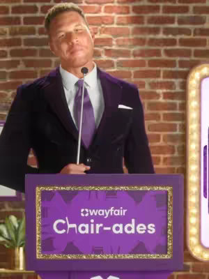 A man in a suit stands behind a purple podium that has the Wayfair logo and the words "Chair-ades" on it. The background is a brick wall, and there's a golden-framed light to the right of the man.