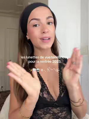A woman with long brown hair, wearing a black headband and a black lace top, is in the center of the image. The text overlay reads, "les lunettes de vue tendances pour la rentrée 2025,", along with a few design elements in the center, which are star, lines, a stick figure, and a flower. She's looking towards the camera with her hands partially raised.