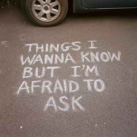 The image shows chalk-written text on asphalt. The words, in white chalk, read: THINGS I WANNA KNOW BUT I'M AFRAID TO ASK. In the background, a car with a tire is partially visible.