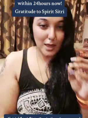 A woman is centered in a video cover image, appearing to be in a home setting. She has long dark hair, a fair complexion, and is wearing a black top with a white patterned design. Overlay text in a navy blue banner reads, "They will call/text within 24hours now! Gratitude to Spirit Sitri". The background features a curtain and indistinct objects, suggesting an intimate filming location. Her expression is gentle, and she appears to be communicating directly to the viewer. The composition is a medium shot emphasizing the upper body and includes her gesturing with her right hand.