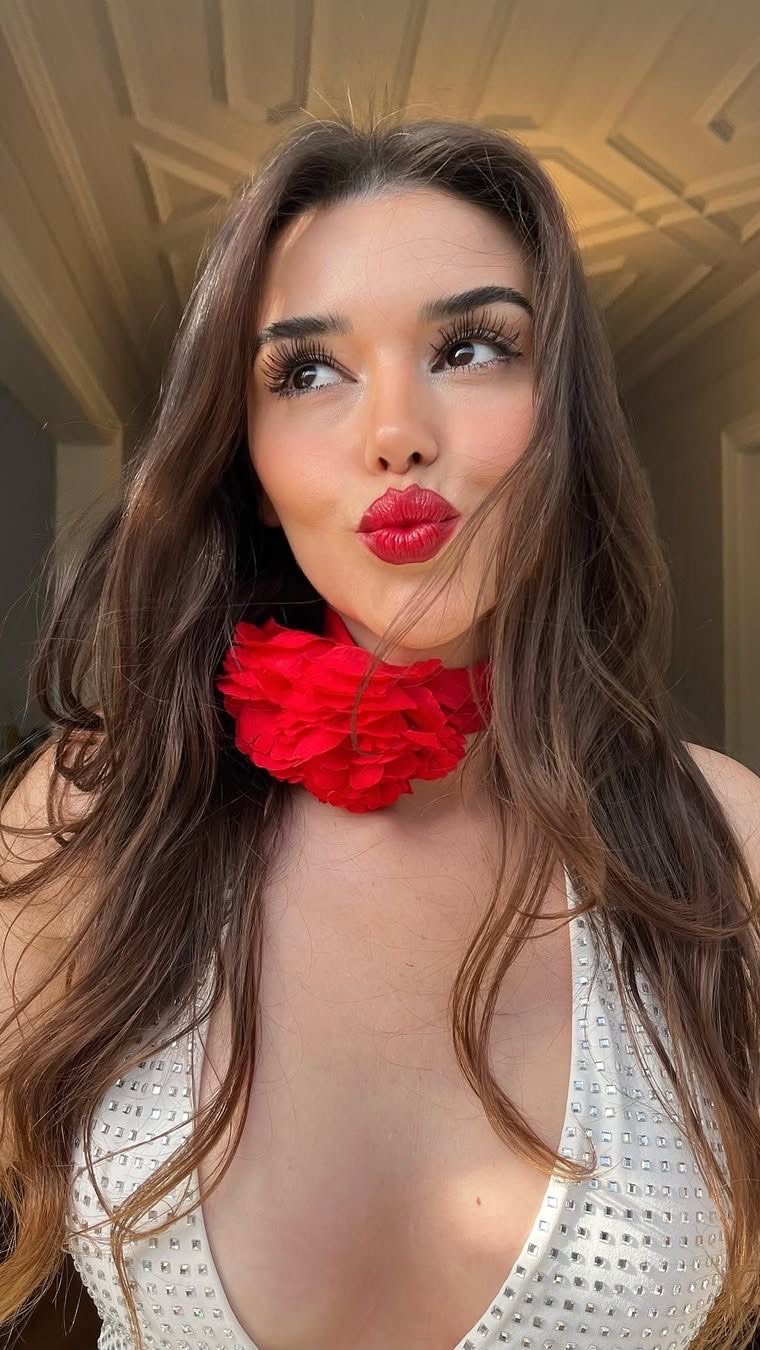 A young woman with long brown hair, bright red lipstick, and long eyelashes is seen puckering her lips. She wears a white top with a bedazzled design. A red flower collar is around her neck. The background is slightly blurred showcasing a room lit with warm light.