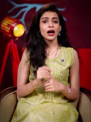 A woman with long dark hair is centered in the shot. She wears a light green sleeveless dress, a necklace and earrings and is seated in a chair. The background features a mix of dark and reddish hues with reddish highlights. To the left is a red tripod with stage lighting.