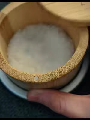 The image is a close-up shot of a wooden container holding white rice, partially concealed by a lid. A hand is visible in the foreground, interacting with the container. The color scheme is dominated by warm tones, contrasting the white rice within the wooden container.