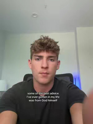 A young man with light skin and short, tousled brown hair is centered in the frame, gazing towards the camera with a neutral expression. He wears a dark t-shirt, and the background consists of a pale wall and a glimpse of what appears to be a window on the left. Subtitles are present, containing words of encouragement.
