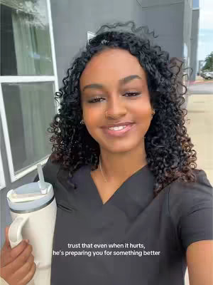 A young woman with curly black hair and a warm smile poses for the camera, holding a white travel mug. She's dressed in a black top. The background shows a building exterior with a window and a hint of a sunny outdoor scene.