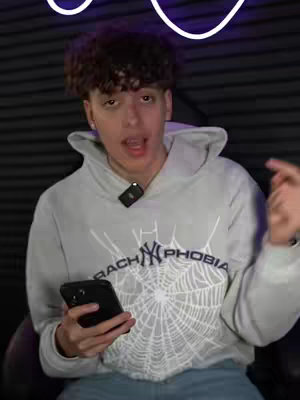 A young man with curly brown hair is centered in a medium shot. He's wearing a light grey hoodie with a black spider web graphic on it, holding a black smartphone in his left hand. A small microphone is clipped on his hoodie near the collar. His mouth is open, and he's pointing to the right with his right hand. The background is a dark gray.