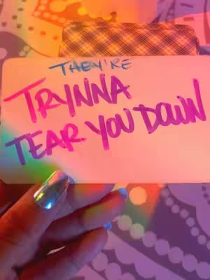 A hand with a long, shiny acrylic nail holds a white card with handwritten text on it. The words "They're Trynna Tear You Down" in neon pink are the focus.