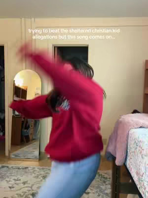 A woman dressed in a red hoodie and light blue jeans is dancing energetically in a room. Her arms are raised, and her body is blurred due to movement. The background shows a bedroom with a mirror, a bed, and other furniture, suggesting a casual, intimate setting.