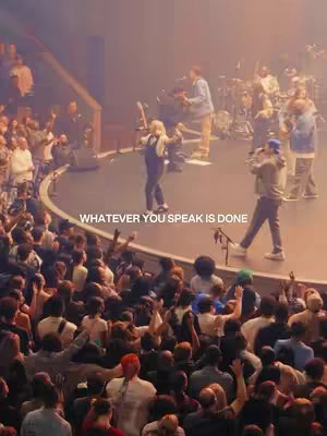 A wide shot captures a stage performance at a church during what seems to be a worship service or concert. A band is playing on a circular stage, with the audience in tiered seating, focused on a group of performers, including singers holding microphones and musicians. Text overlayed on the image says “WHATEVER YOU SPEAK IS DONE”. The scene is lit with warm tones.