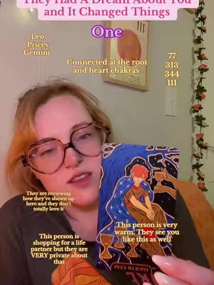 A vertical shot with the creator, a person with light skin and blonde hair wearing glasses, holding a tarot card. The card features a man with red hair, covered in a blue cloak, with a purple background, and holding three cups. The creator's face is somewhat obscured, she has a neutral expression. Text overlays appear around her, including astrological signs and phrases.