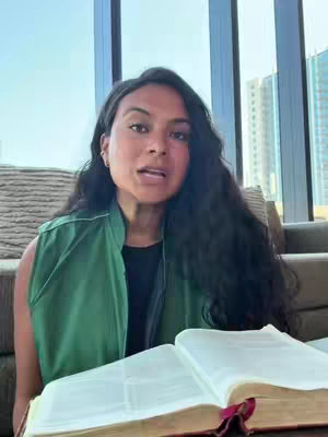 A woman with long dark hair and a green vest sits in front of a bright window. She holds an open book, likely a Bible, with a red ribbon bookmark.