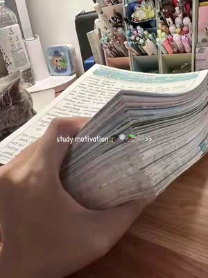 The image shows a hand holding a thick stack of papers, likely a textbook or notebook. The foreground is dominated by the hand and papers, while the background has a desk, a shelf full of pens, a laptop, and a grey cat. The text 'study motivation' is overlaid on the image.