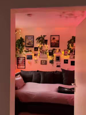 The view into a bedroom shows a cozy, well-lit space. The foreground is the doorway, which is brown. Inside, the room has a pinkish/red glow from ambient lighting. Pictures and decorations adorn the wall, arranged in a gallery style, and the focus is on the bed and surrounding elements.