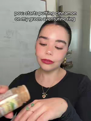 A woman in a black top holding a cinnamon shaker and showing off a green aventurine ring, against a blurred white background. Text overlay says "pov: starts putting cinnamon on my green aventurine ring". 