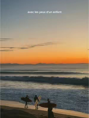 A scenic beach at sunset with surfers. The sky transitions from a bright orange near the horizon to a lighter blue at the top. Three surfers are silhouetted, walking on the shoreline holding surfboards. Gentle waves roll onto the beach.