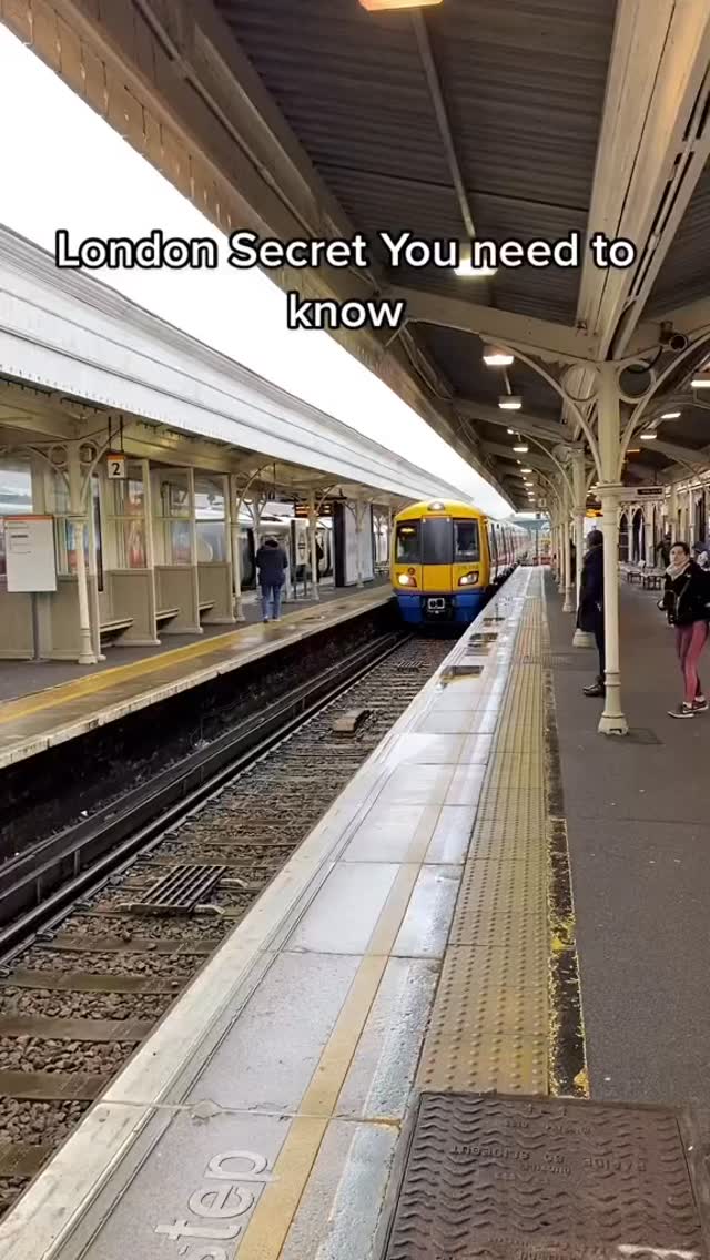 A shot from inside a train station in London. A yellow and blue train pulls into the station, with details like the platform and overhead structure visible. People are present, some waiting for the train and others walking by. The lighting is bright, indicating daylight.