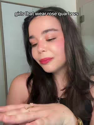 A woman with long dark hair, wearing a black tank top, and makeup including pink blush and red lipstick. She is holding her left hand up, displaying a ring, and there is a necklace visible. The background is a light-colored wall and door.