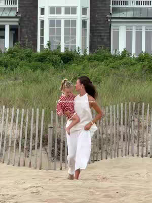 A woman in a white dress and a young child in a striped shirt are walking on a sandy beach in front of a wooden fence and tall grass. Behind them are a tall gray building and green foliage, showcasing a bright, sunny day.