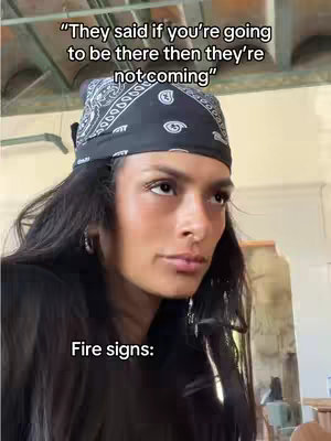 A medium shot features a young woman with long, dark hair and a black bandana. Above her, text overlaid on the image reads, 'They said if you're going to be there then they're not coming.' Below her, the text 'Fire signs:' is displayed. The background appears to be an interior setting with warm lighting.