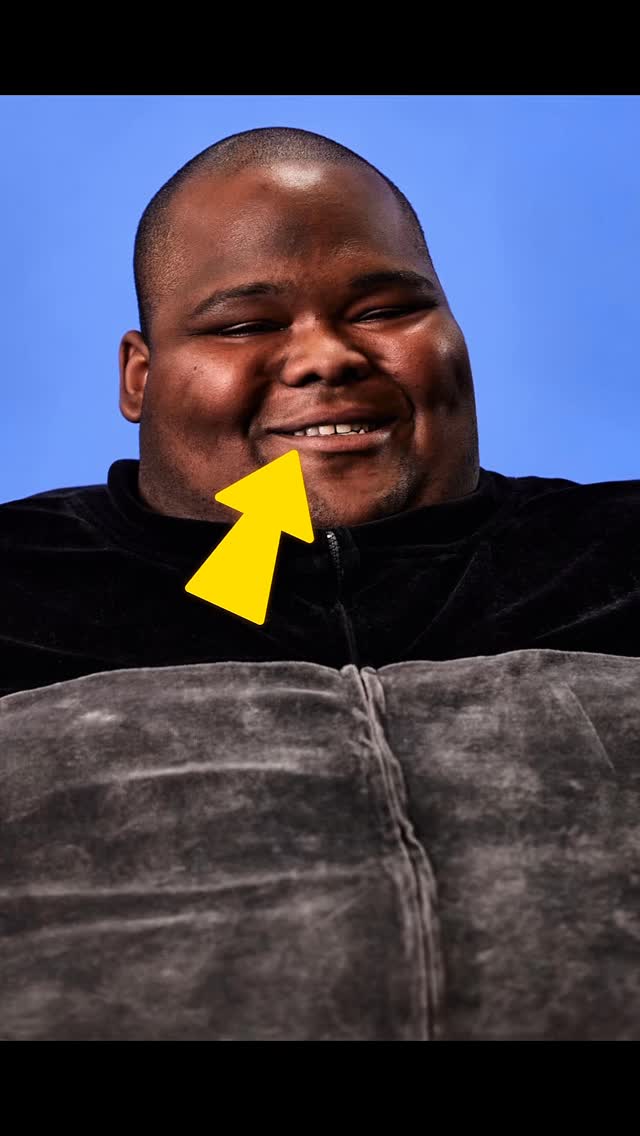 The image captures a close-up of a man's face with a blue background. The man, who appears to be of African descent, is smiling, revealing his upper teeth. A bright yellow arrow points towards his mouth. He is seen wearing a black, zippered jacket and the lower portion of a greyish-brown seat.