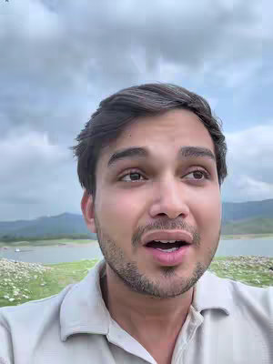 A selfie-style shot of a man looking upwards with an open mouth, set against a backdrop of a cloudy sky, a lake, and distant mountains. The man has short, dark hair, a trimmed beard, and is wearing a light-colored collared shirt. The overall composition has a natural, outdoor feel.