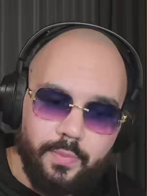 A medium shot features a man in sunglasses and headphones. The man has a shaved head, full beard, and is wearing gold-rimmed, ombre sunglasses. He has a black headphone set around his neck.