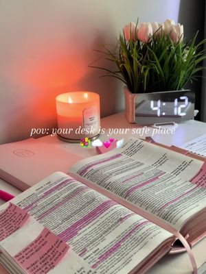 A well-lit, cozy desk scene. A lit candle, a digital clock displaying 4:12, and a small plant with white flowers sit on a desk. An open book with pink highlights and sticky notes, as well as a closed book, are present, suggesting a study or devotional setting. The overall aesthetic is warm and inviting.