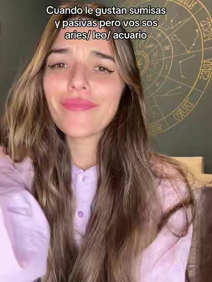 A young woman is centered in the frame, facing the camera with a slight smile. She has long, light brown hair. She is wearing a light purple collared shirt. Behind her, a circular design with astrological symbols is visible on a dark background. The lighting is soft, and the overall composition is simple.