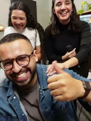 A selfie-style image with three people. In the foreground, a man with glasses and a beard smiles broadly, giving a thumbs-up. Two women are in the background, also smiling and laughing. The composition conveys a sense of joy and camaraderie.