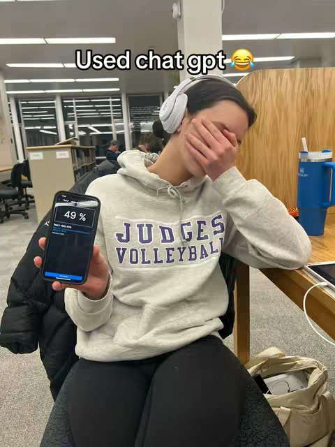 A young woman in a hoodie is holding a phone displaying a poor test score while covering her face with her hand, suggesting embarrassment. She's wearing headphones and sitting in what appears to be a library setting. The text overlay reads 'Used chat gpt'. The background has a wooden panel with the woman in a lightly lit environment.
