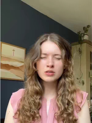 A woman with long, wavy blonde hair is filmed from the chest up. She is wearing a light pink top. Her eyes are closed, and she appears to be singing or in a moment of contemplation. The background includes a wall painted in dark blue with a framed artwork of a cross and a beige cabinet holding plants.
