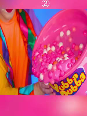 The image shows a person's arms holding a very large, pink container with candy-like items spilling out. The container has a purple and yellow logo at the bottom. The person's arm on the left is wearing a colorful, clown-like outfit, with shades of orange and green, while the rest of the image is bathed in a vibrant pink. The number (2) is at the top of the image.