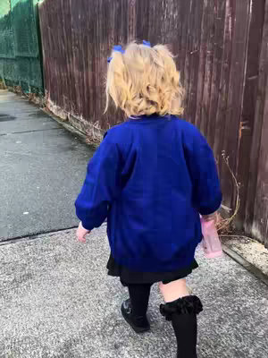 A young girl with blonde pigtails, wearing a blue sweater and black skirt, walks away from the camera on a sidewalk. She carries a pink water bottle. The background consists of a wooden fence and a green fence. The lighting appears to be overcast.