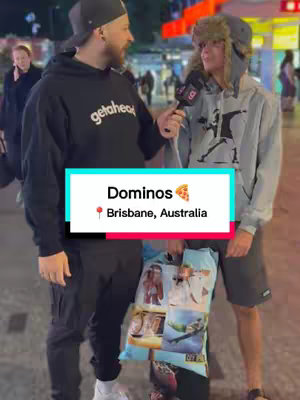 Two young men are standing outdoors, conversing. One is holding a microphone, as if interviewing the other. A banner with the Domino's logo and location is overlaid on the image. The background is blurred, suggesting a busy street scene at nighttime.