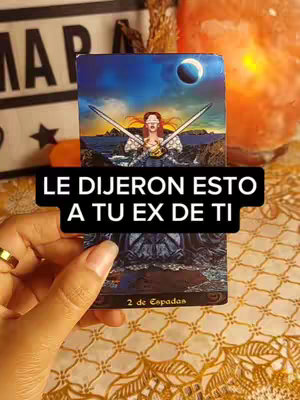 A hand holds up a tarot card from the '2 of Swords' deck, featuring a woman with crossed swords. The card is overlaid with a black text box containing white text. The background has blurred elements of a crystal, amber lighting, and a patterned surface.