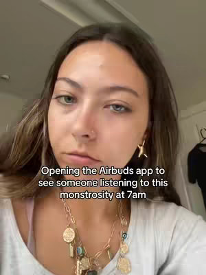 A selfie-style shot featuring a young woman with brown hair and a neutral expression, taken from a close perspective. The text overlay reads, "Opening the Airbuds app to see someone listening to this monstrosity at 7am." She wears a white top, a necklace, and a nose ring, and the background is a simple interior setting.