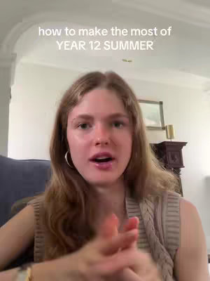 A young woman with fair skin and long, light brown hair is centered in the frame. She's wearing a sleeveless, cream-colored knit top. The background includes a white ceiling with a curved architectural detail at the top, revealing a glimpse of a room behind. Her hands are clasped together in front of her, and she appears to be speaking or giving a presentation. The lighting seems natural, possibly from a window out of frame.