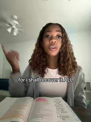A young woman with curly hair, wearing a gray sweater, is sitting in a room. She is holding an open book with the left hand and pointing with index finger, while looking directly at the camera. Text overlayed in the image.