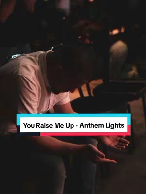 A man wearing a light-colored shirt is seen from the chest up, in a slightly darkened setting. He is shown from the side and is kneeling down, with his head bowed and hands extended outward, palms up, as though in a gesture of supplication or offering. The background is blurred, suggesting a focus on the man. A text overlay highlights the song title and artist across the image's lower portion.