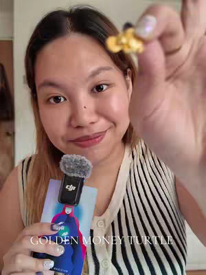 A selfie-style shot featuring a fair-skinned woman holding a small microphone with a fuzzy windscreen. She holds a gold-colored object in her other hand towards the camera, possibly a charm or small figurine. The background is slightly blurred, but one can see the woman’s casual attire and striped top, and a card. The focus is on the woman’s face and upper body.