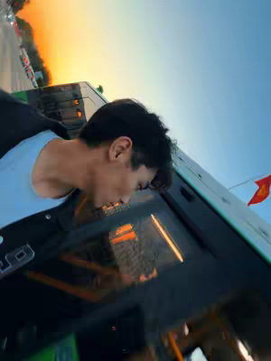 A man leans out the window of a bus with a sunset in the background, the bus's window and the person's silhouette are reflected in the sky.
