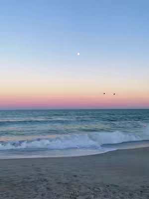 The image depicts a serene beach scene at dusk or dawn. The sky transitions from a light blue at the top to pink and orange towards the horizon. The ocean is a deep teal, with small waves gently rolling onto the sandy beach. Two dark shapes, likely birds, are visible in the sky near the horizon, and a bright, round object, perhaps the moon or sun, is centered above the horizon. The overall composition is peaceful and calming.