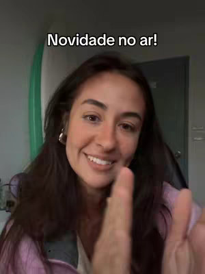 A young woman with long dark hair is smiling directly at the camera. The text overlay says, "Novidade no ar!" (News in the air!). She is wearing large hoop earrings, and a pink top. Her right arm is raised, suggesting emphasis. A surfboard and green wall are slightly visible in the background.