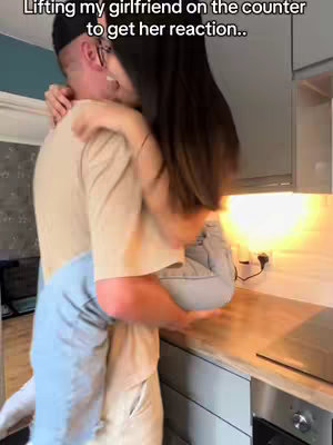 A man in a beige shirt and a black cap is lifting a woman with long black hair. They are in a kitchen setting, with a wooden countertop, stainless steel appliances, and a bright overhead light. The woman is wearing light blue jeans, and the man is wearing a light-colored t-shirt and glasses. The man has his arms around the woman’s waist holding her up, and the woman is leaning into the man’s shoulder.