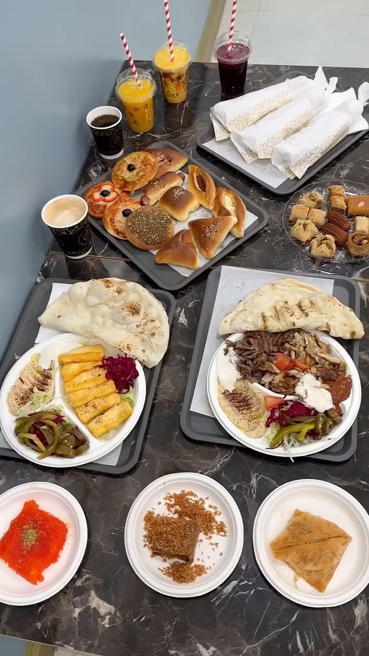 A top-down shot showcases a table laden with food and drinks. Several dishes are displayed on trays, including plates of shawarma, falafel, and fries, along with assorted pastries and baked goods. Various drinks, like orange juice and coffee, are arranged around the food. The composition shows a variety of colors and textures, emphasizing the food's presentation.
