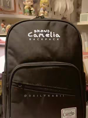 The cover image shows a close-up of a black backpack with the brand name 'Camelia Backpack' in white, stylized text. Below the brand name is the text '@DAILYNARII' in a similar white font. In the background are a few figurines on a shelf. The composition centers on the backpack, suggesting a review or unboxing video.